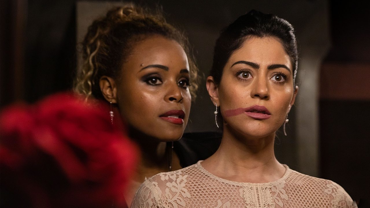 Duas mulheres em cena dramática; uma encara à frente com expressão tensa e marca de batom no rosto, enquanto a outra a observa de perto com olhar intenso