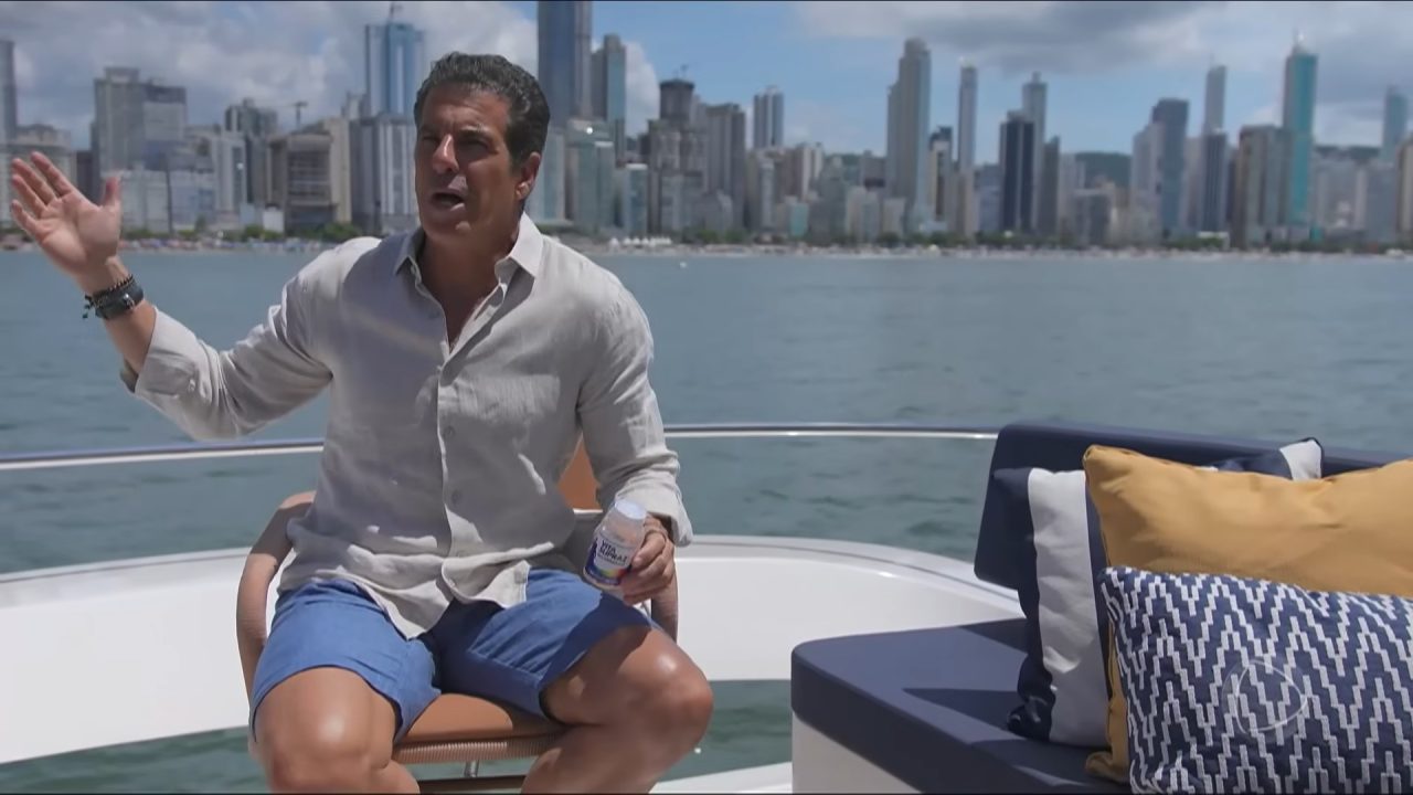 Alvaro Garnero, apresentador do 50 por 1, gesticula sentado em barco com skyline de Balneário Camboriú ao fundo durante gravação do Domingo Espetacular.