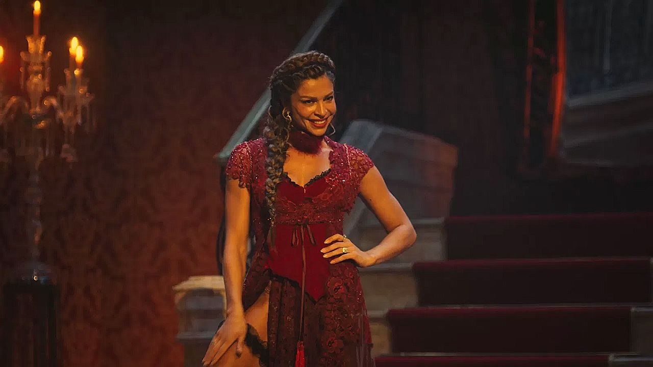 Mulher veste figurino vermelho com renda e posa em cenário teatral iluminado por luz quente. Ela sorri com expressão confiante, com uma das mãos na cintura, diante de escadaria e candelabros acesos, sugerindo apresentação em palco ou espetáculo musical.