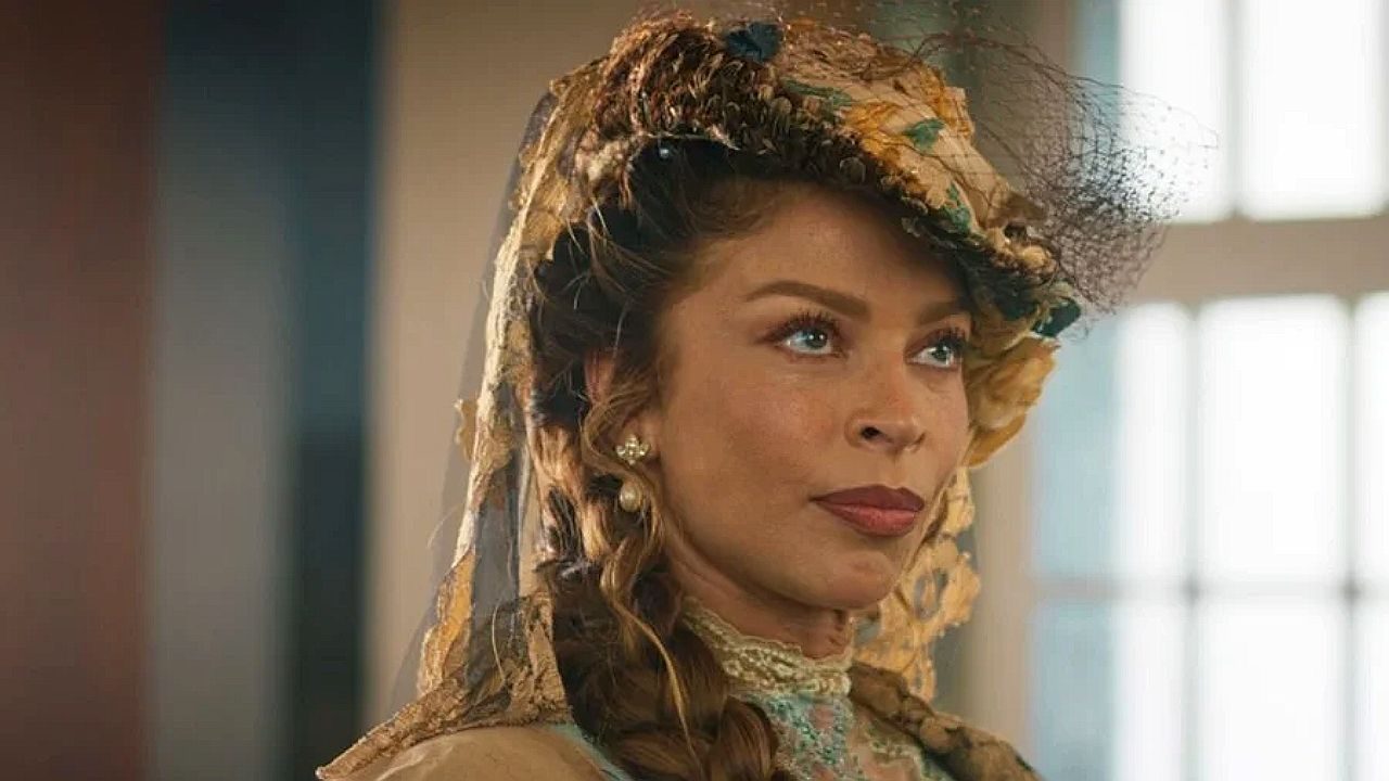 Grazi Massafera em Dona Beja aparece em um ambiente interno, usando roupas de época elegantes, incluindo um vestido detalhado e um chapéu ornamentado com véu. Seu cabelo está estilizado de forma elaborada, com tranças e cachos, e ela usa brincos delicados. A expressão é séria e composta, olhando levemente para o lado. A iluminação suave e o figurino sugerem uma cena ambientada em período histórico ou produção dramática.