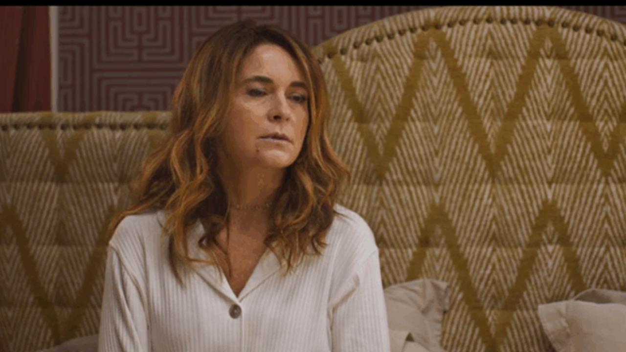 Mulher sentada em uma cama com expressão séria, vestindo roupa clara, em quarto com cabeceira acolchoada de padrão geométrico e tons terrosos