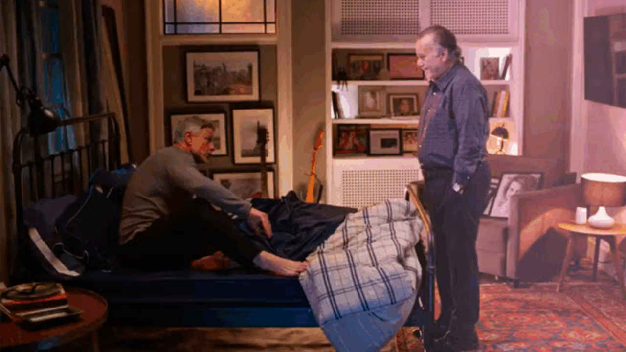 Cena da novela Dona de Mim mostra dois homens em um quarto aconchegante — um sentado na cama, o outro em pé — em meio a uma decoração com quadros, livros e luz ambiente amarelada