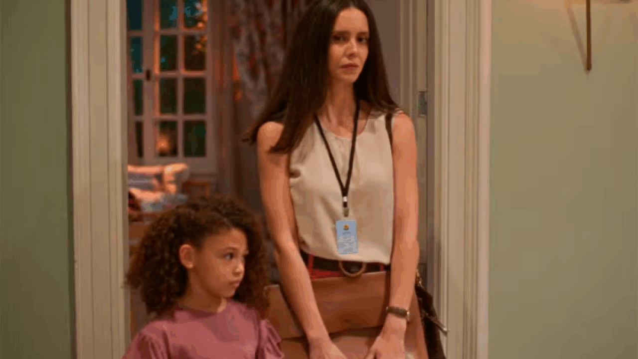 Mulher com expressão séria, vestindo blusa clara e crachá no pescoço, está ao lado de uma menina de camiseta rosa em uma porta de casa, observando algo no interior do cômodo iluminado.