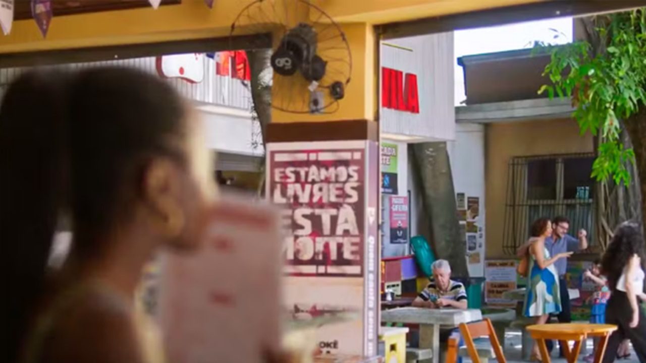 Ambiente de bar ou espaço cultural aberto com mesas de madeira e cadeiras espalhadas, em primeiro plano à esquerda aparece uma mulher desfocada segurando um copo próximo ao rosto com o fundo em foco, ao centro há um cartaz fixado em uma coluna com os dizeres “Estamos Livres Esta Noite” em letras grandes e claras, acima do cartaz um ventilador de parede antigo preso à coluna, ao fundo vê-se uma área externa com pessoas conversando em pé e sentadas, incluindo um homem idoso sentado sozinho à mesa e um casal em pé conversando próximo a um balcão, prédios simples ao redor com fachadas desgastadas e placas comerciais visíveis, iluminação natural diurna cria um clima cotidiano e urbano de bairro popular ou centro cultural informal