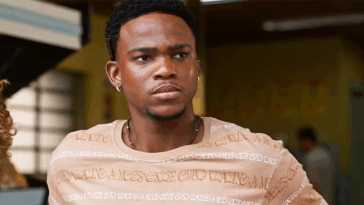 Homem com expressão séria e olhar determinado aparece em ambiente interno, vestindo camiseta bege estampada e colar simples, sugerindo um momento de tensão ou reflexão