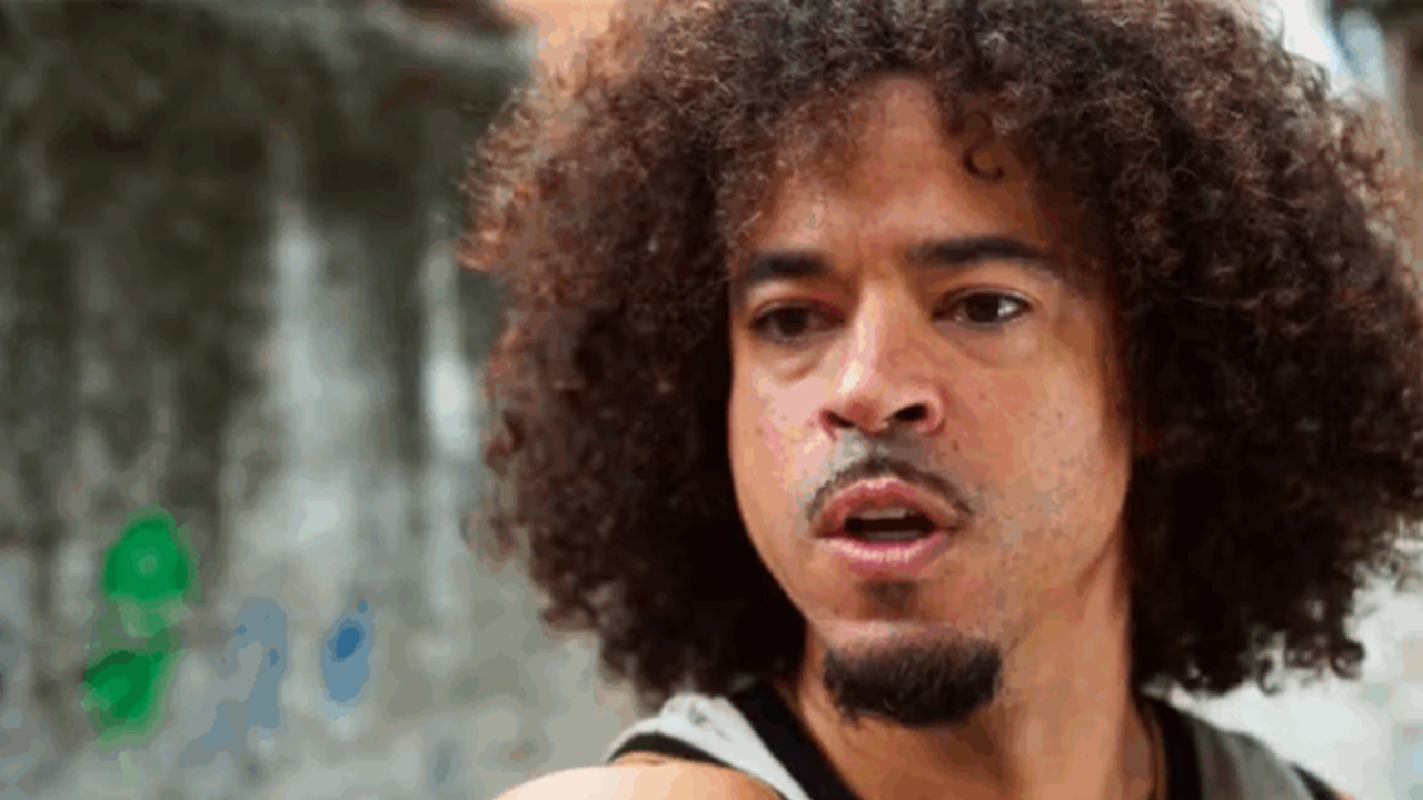 A imagem mostra um homem com expressão séria, olhando para o lado, em um ambiente externo. Ele tem cabelos cacheados e volumosos, barba rala e está vestindo uma regata cinza. Ao fundo, há uma parede desfocada com tons de cinza e algumas marcas coloridas