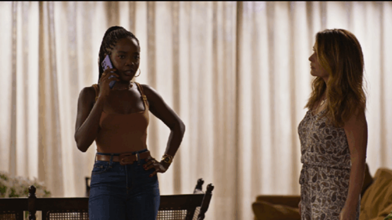 A imagem mostra duas mulheres em uma sala iluminada por luz suave, com cortinas claras ao fundo. À esquerda, uma mulher está ao telefone, com expressão séria, usando blusa marrom e calça jeans. À direita, outra mulher a observa atentamente, vestindo um macacão estampado