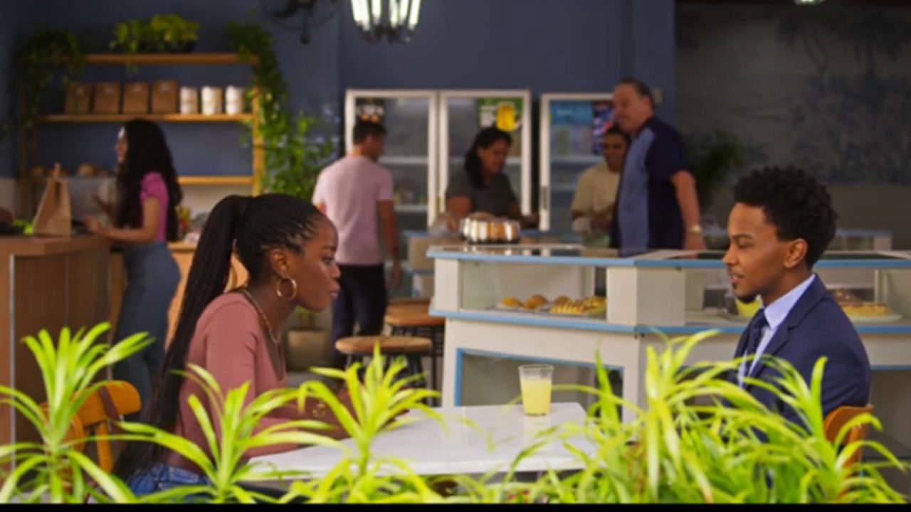 Mulher e homem sentados frente a frente em uma mesa de lanchonete iluminada, com plantas em primeiro plano e um copo de suco amarelo sobre a mesa. A mulher usa blusa rosa e brincos de argola, enquanto o homem veste terno azul. Ao fundo, há pessoas atendendo e conversando no balcão, prateleiras com produtos e uma decoração em tons de azul e madeira.