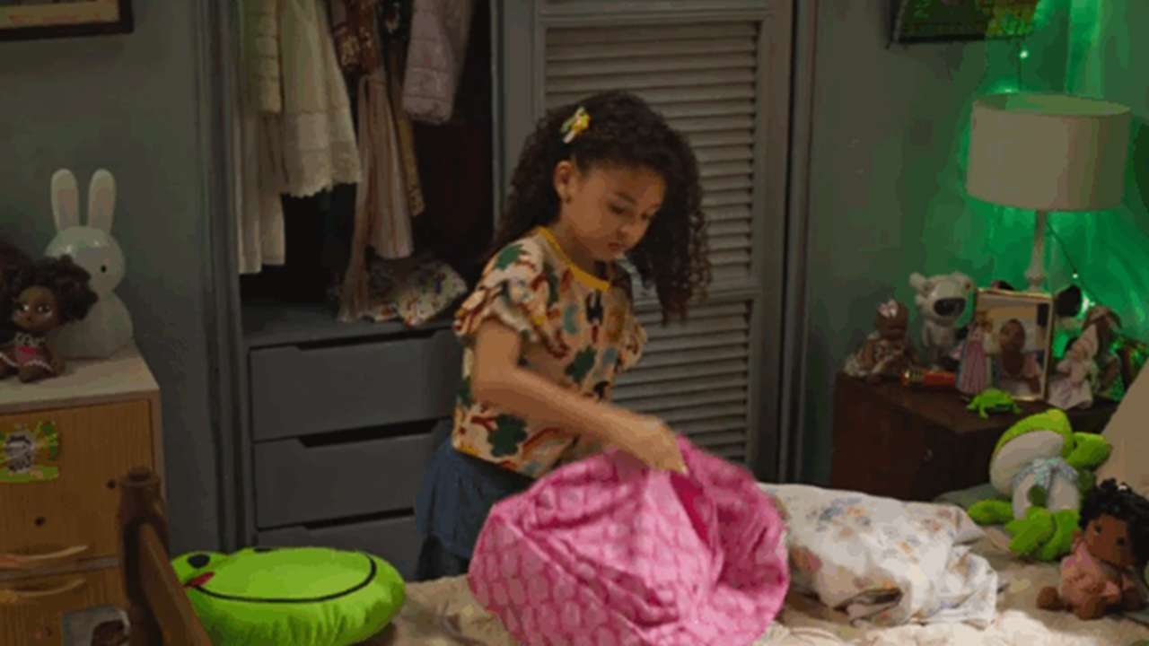 Cena da novela Dona de Mim, mostrando a personagem infantil Sofia em seu quarto, arrumando uma mochila cor-de-rosa sobre a cama, cercada por brinquedos e objetos coloridos