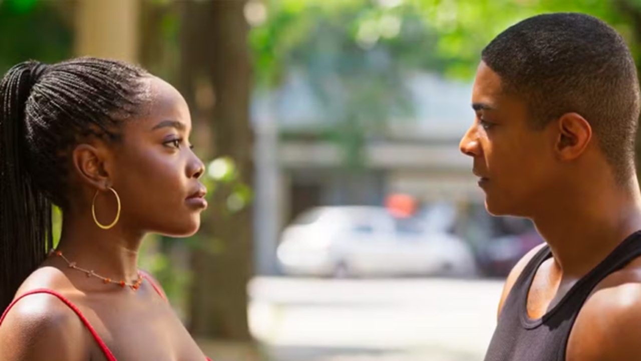 Duas pessoas se encaram em uma rua ensolarada, uma mulher de cabelos trançados presos em rabo de cavalo usa vestido vermelho e brincos de argola dourados, enquanto o homem à frente veste regata preta; o ambiente ao fundo mostra árvores e carros desfocados.