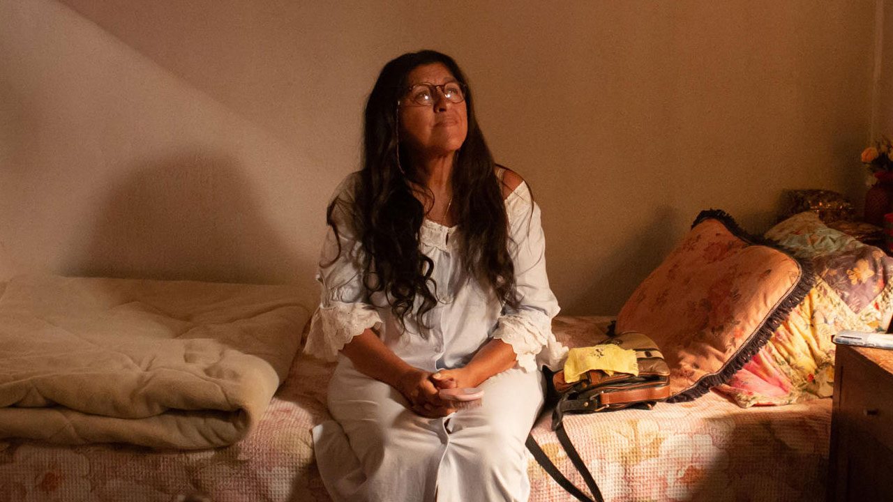 Dona Lurdes (Regina Casé) sentada na beira da cama com expressão emocionada
