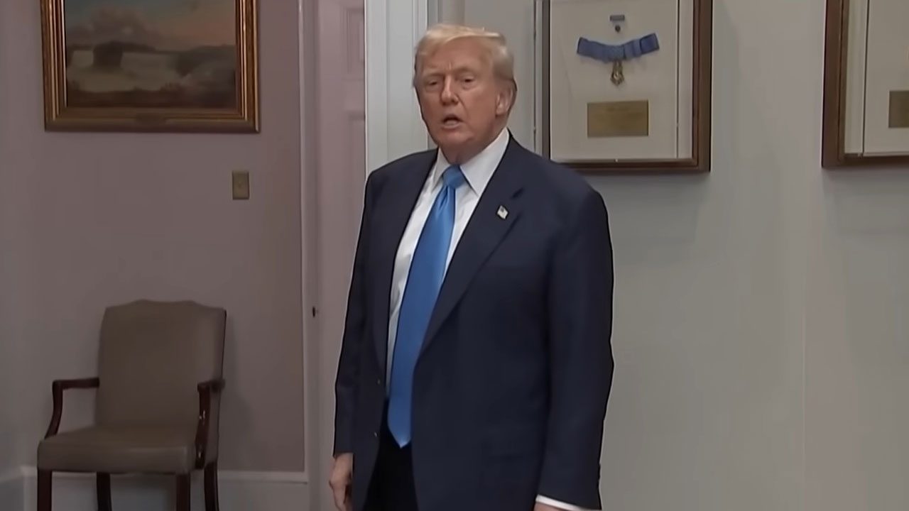 Donald Trump de terno escuro e gravata azul em pé dentro de sala com quadros e medalhas na parede