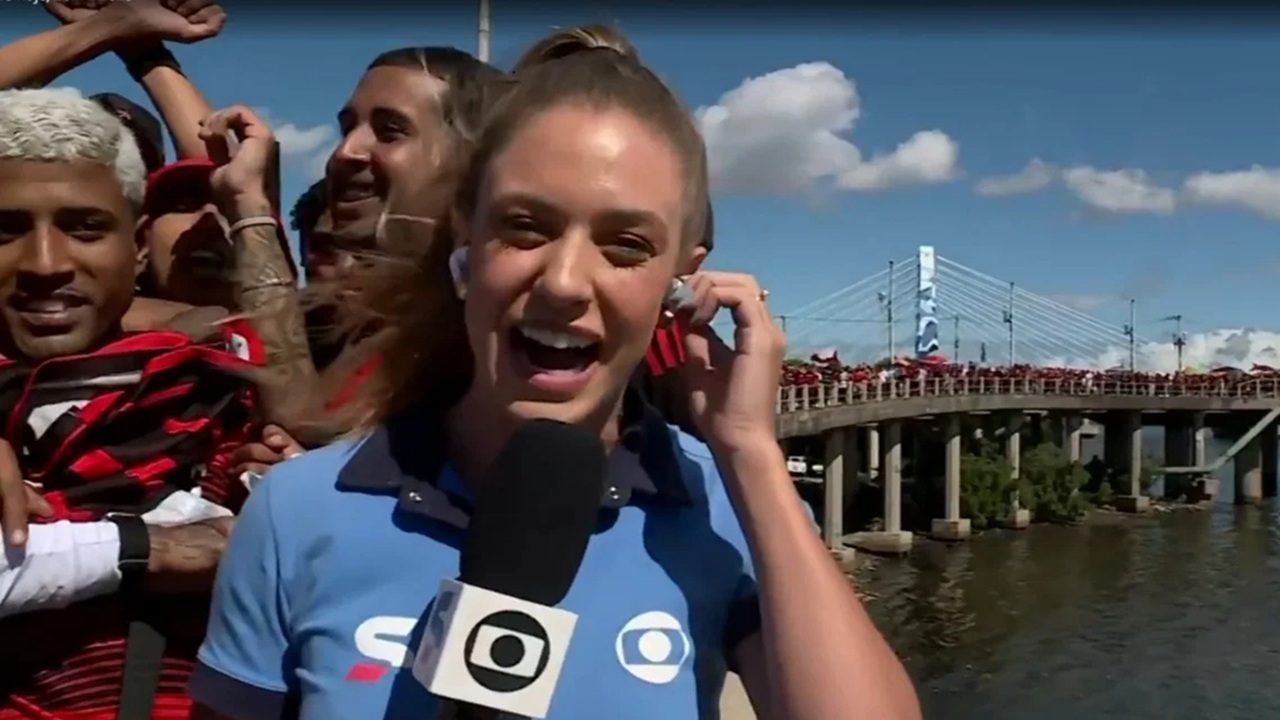 Repórter sorrindo com microfone da Globo durante cobertura de torcida em ponte lotada ao fundo