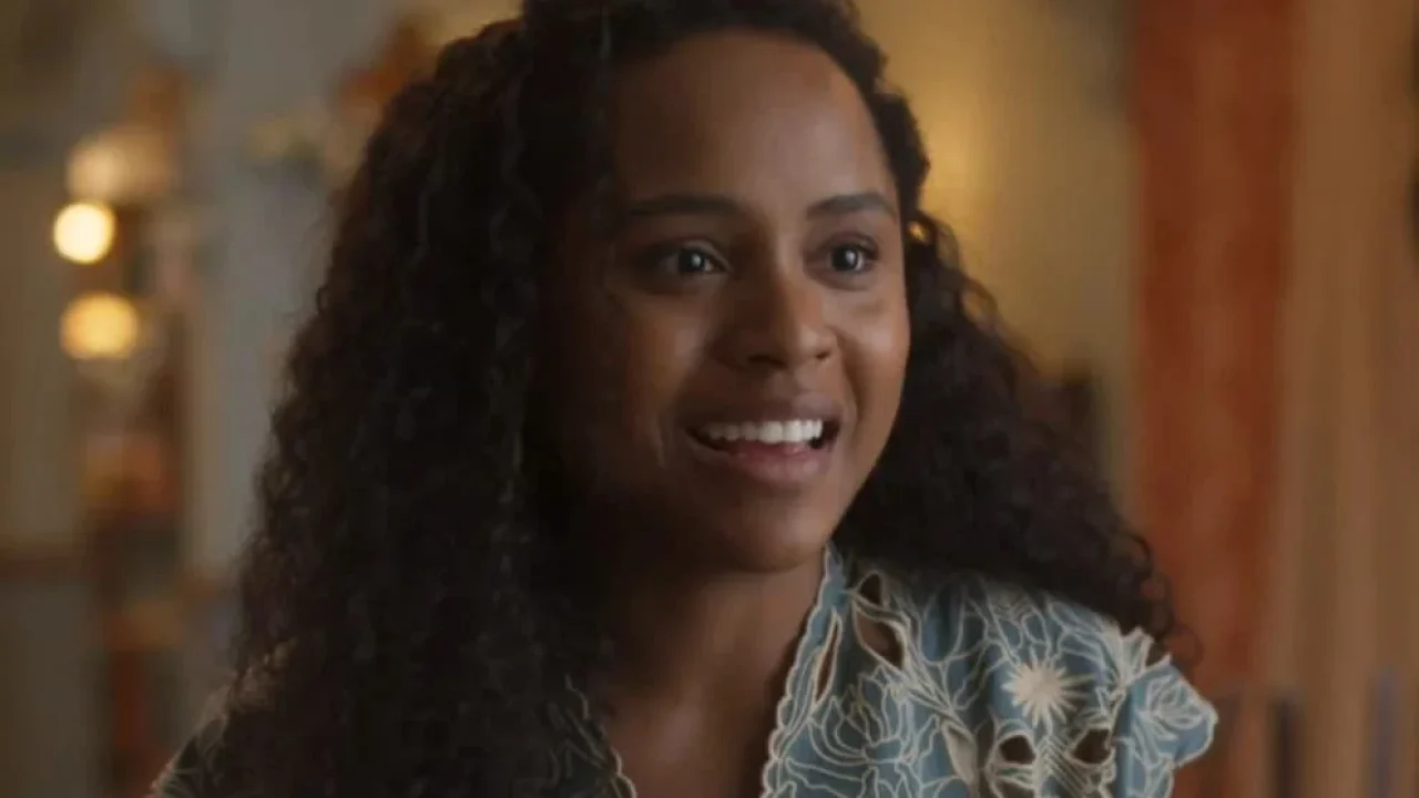Duda Santos sorri em cena de novela, usando blusa estampada e iluminação suave ao fundo