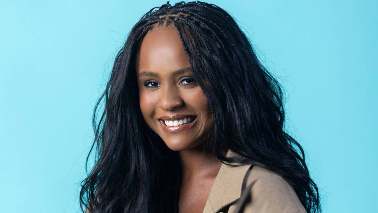 Duda Santos sorri em foto de estúdio com fundo azul usando casaco bege e cabelo solto com tranças na parte frontal