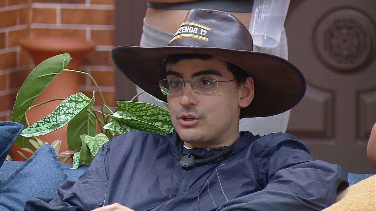Dudu Camargo com jaqueta azul, óculos e chapéu de cowboy com o logo A Fazenda 17 conversa na área interna do reality show da Record