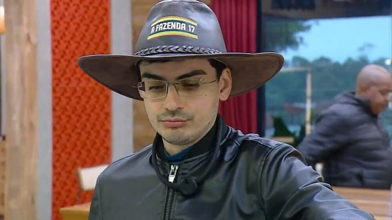 Homem de óculos com chapéu de A Fazenda 17 e jaqueta preta em área interna do reality show