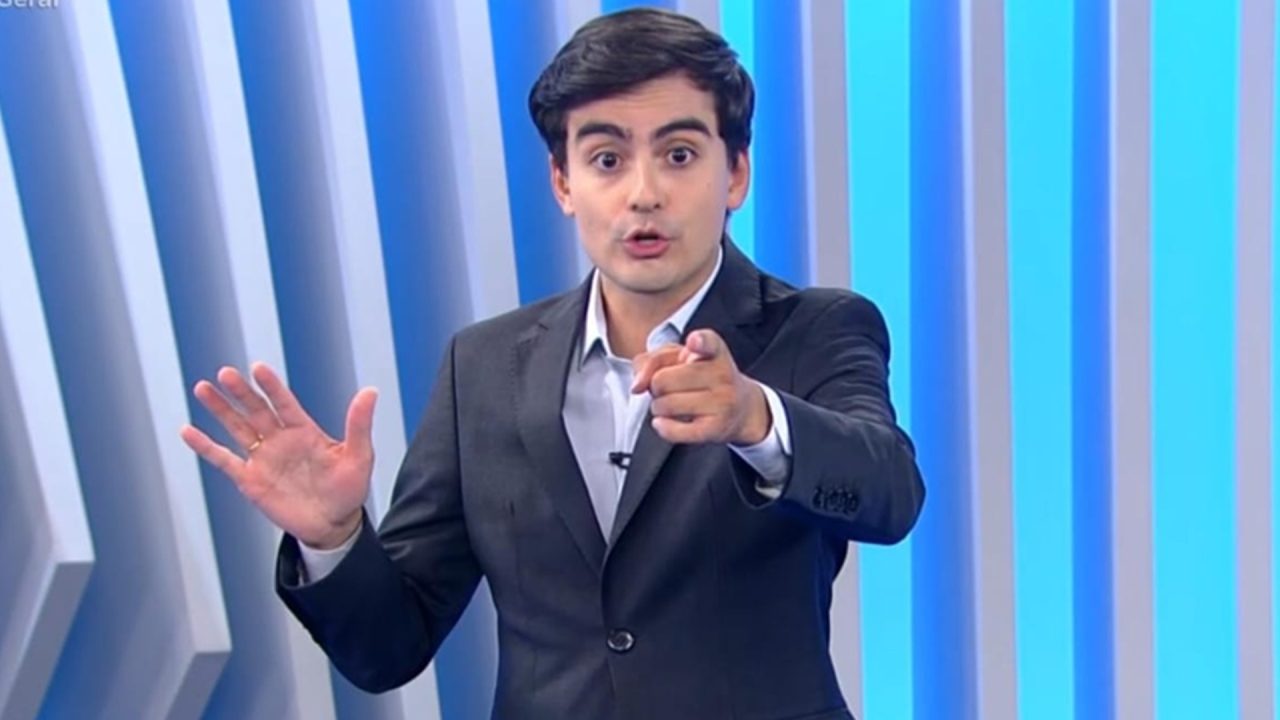 Dudu Camargo, de terno escuro e camisa clara, fala no estúdio do Balanço Geral, com fundo em tons de azul, apontando o dedo para a câmera enquanto mantém a outra mão levantada, em gesto enfático.
