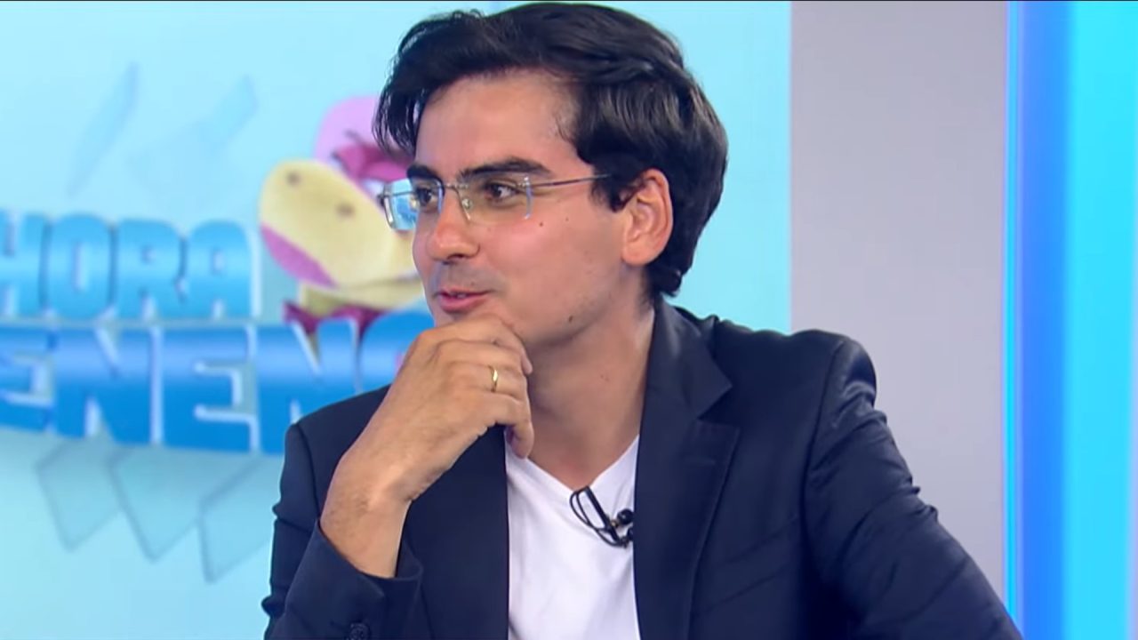 Dudu Camargo, um homem de óculos e cabelo escuro, participa do Balanço Geral, apoiando a mão no queixo enquanto olha para a lateral; ele veste blazer escuro sobre camiseta branca e usa microfone de lapela, com painel colorido ao fundo indicando cenário de estúdio.