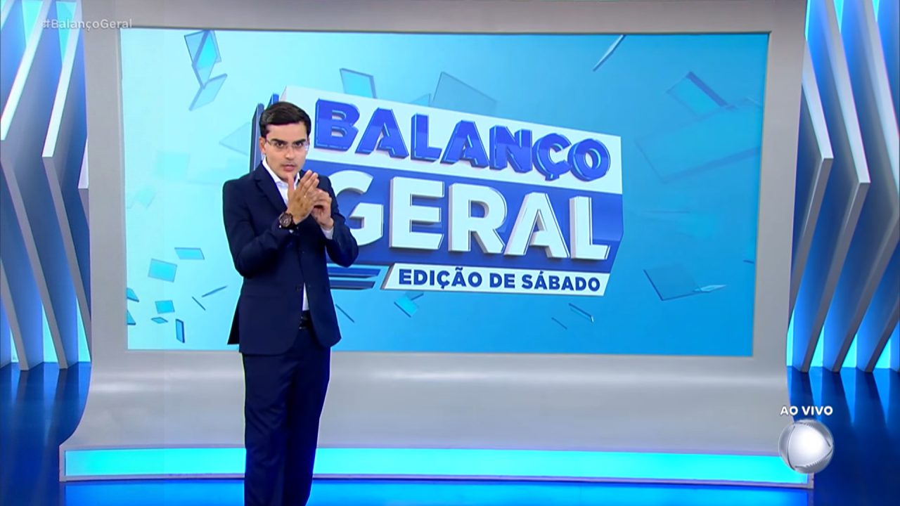 Dudu Camargo, de terno escuro, está em pé no estúdio diante de um grande telão com o título Balanço Geral – Edição de Sábado; no canto da tela aparece a indicação ao vivo e o logotipo da Record.