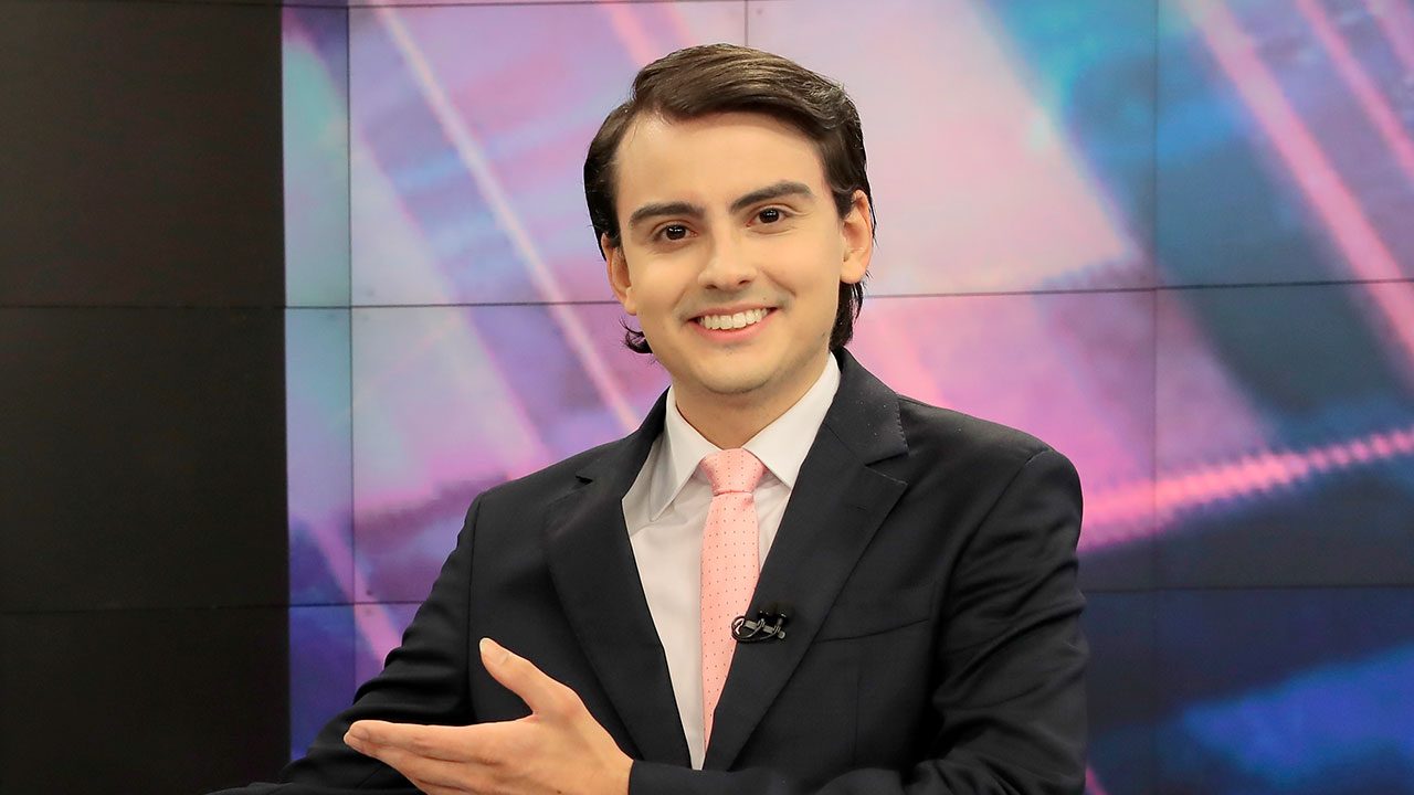 Dudu Camargo sorrindo de terno e gravata rosa no estúdio do Primeiro Impacto no SBT