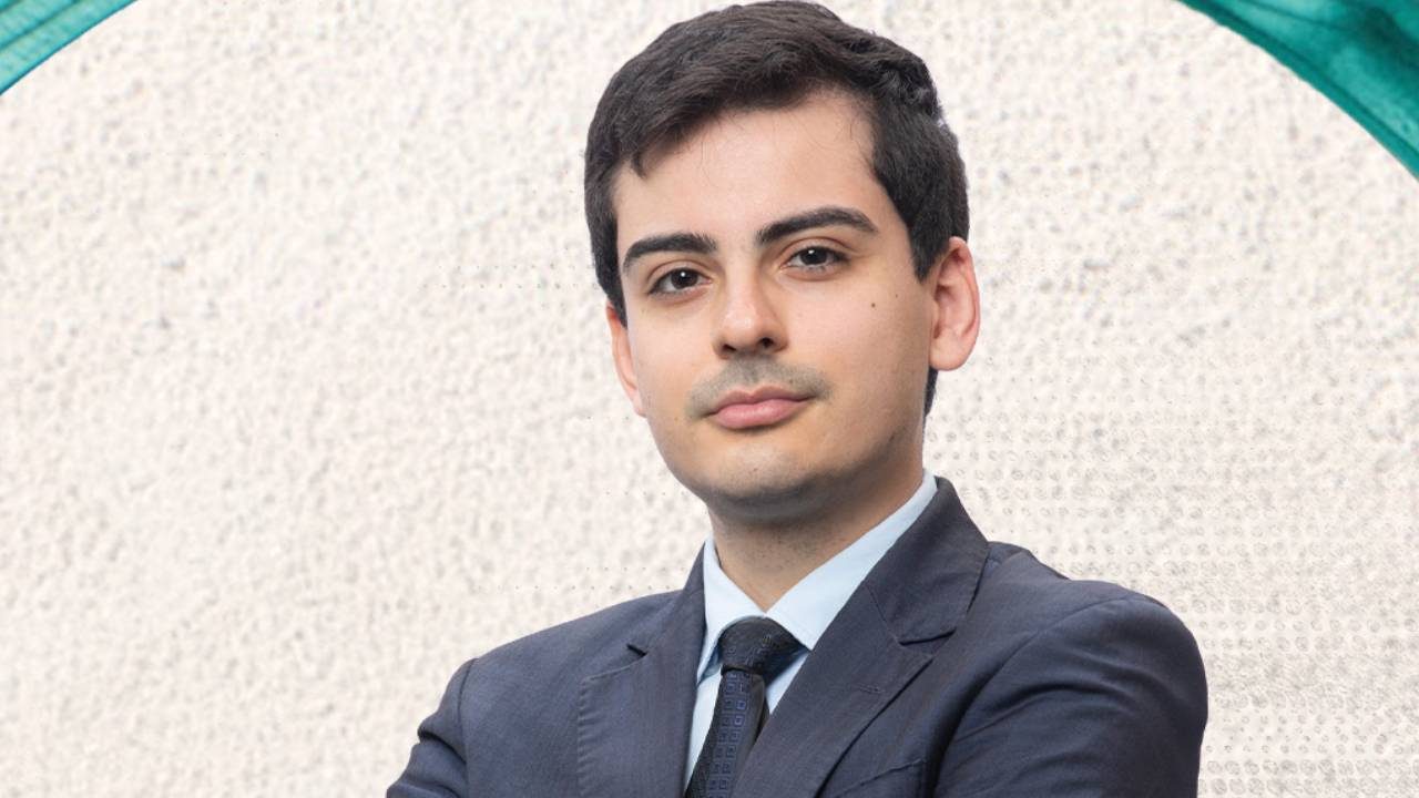 Dudu Camargo de terno escuro e gravata posa com braços cruzados em frente a fundo claro