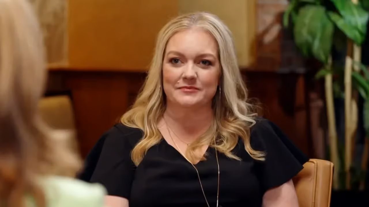 Mulher loira de cabelos ondulados usa blusa preta e observa interlocutora durante conversa em ambiente interno com iluminação quente e plantas ao fundo