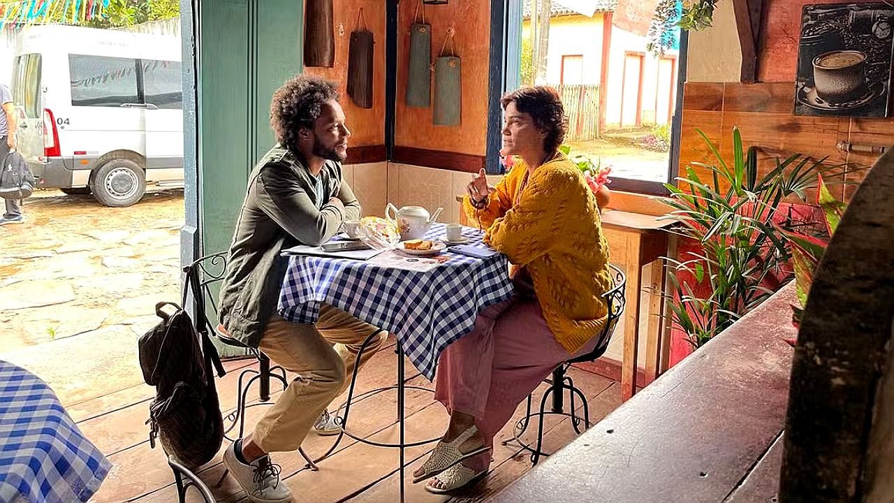 Homem e mulher conversam à mesa em café simples com toalha xadrez e xícaras durante encontro em cidade do interior