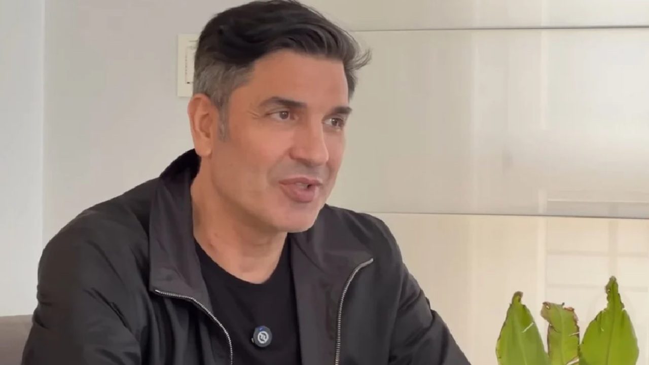 Edu Guedes durante gravação em ambiente interno bem iluminado, usando jaqueta preta e camiseta escura, com uma planta verde em primeiro plano