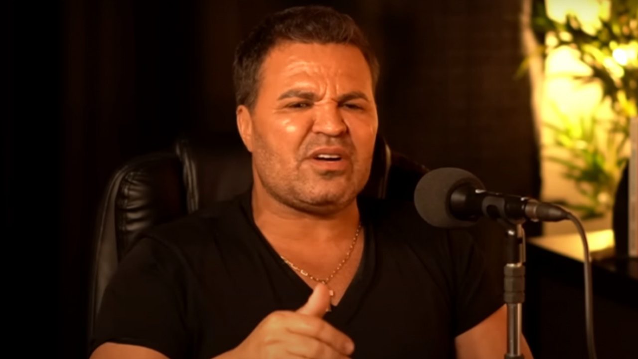 Homem sentado em cadeira preta participa de uma gravação em estúdio, falando diante de um microfone profissional. Ele veste camiseta preta, faz uma expressão de incômodo ou indignação e gesticula com a mão. O fundo é escuro, com iluminação quente e uma planta ao lado