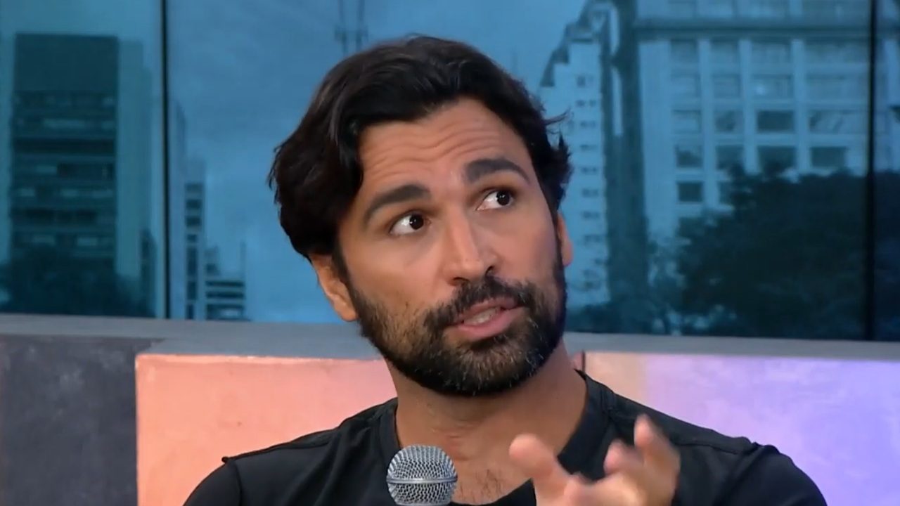 A imagem mostra um homem de barba e cabelos escuros, segurando um microfone e fazendo um gesto com a mão enquanto fala. Ele está em um estúdio de TV com janelas ao fundo que mostram prédios urbanos, sugerindo uma entrevista ou participação em um programa