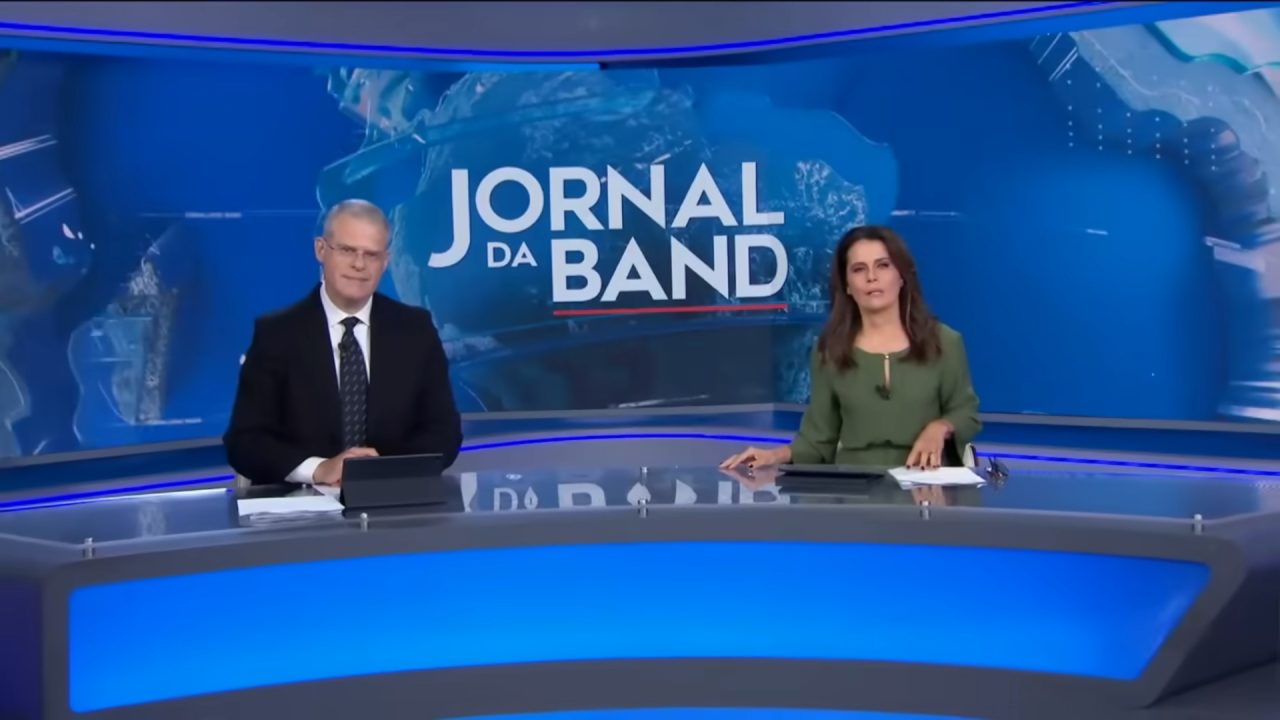 Eduardo Oinegue e Adriana Araújo, apresentadores do Jornal da Band, sentados na bancada durante telejornal com telão azul ao fundo exibindo logotipo do programa.