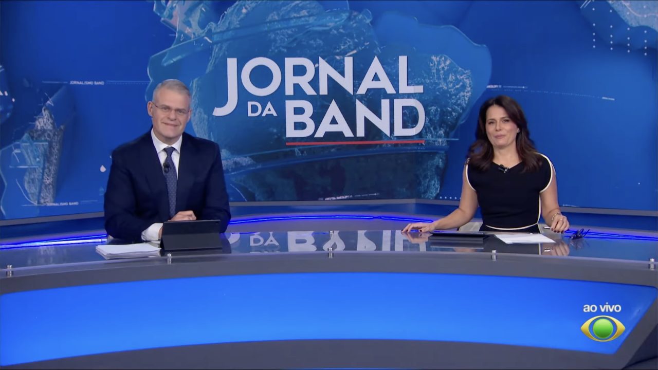 Eduardo Oinegue e Adriana Araújo sorriem na bancada do Jornal da Band, com painel do telejornal ao fundo