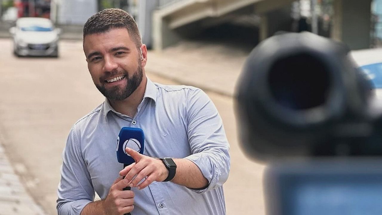 Eduardo Pinzon aparece em uma rua, segurando um microfone e sorrindo enquanto aponta em direção à câmera. Ele usa camisa clara de manga longa, tem barba bem aparada e cabelo curto. À sua frente, há uma câmera profissional desfocada, indicando que ele está sendo filmado. O cenário urbano ao fundo mostra carros e construções, sugerindo uma reportagem externa ou gravação jornalística.