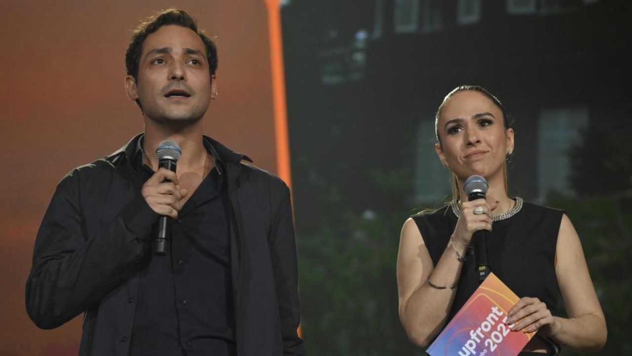 Eduardo Sterblitch e Tatá Werneck seguram microfones durante apresentação em palco de evento com iluminação ao fundo