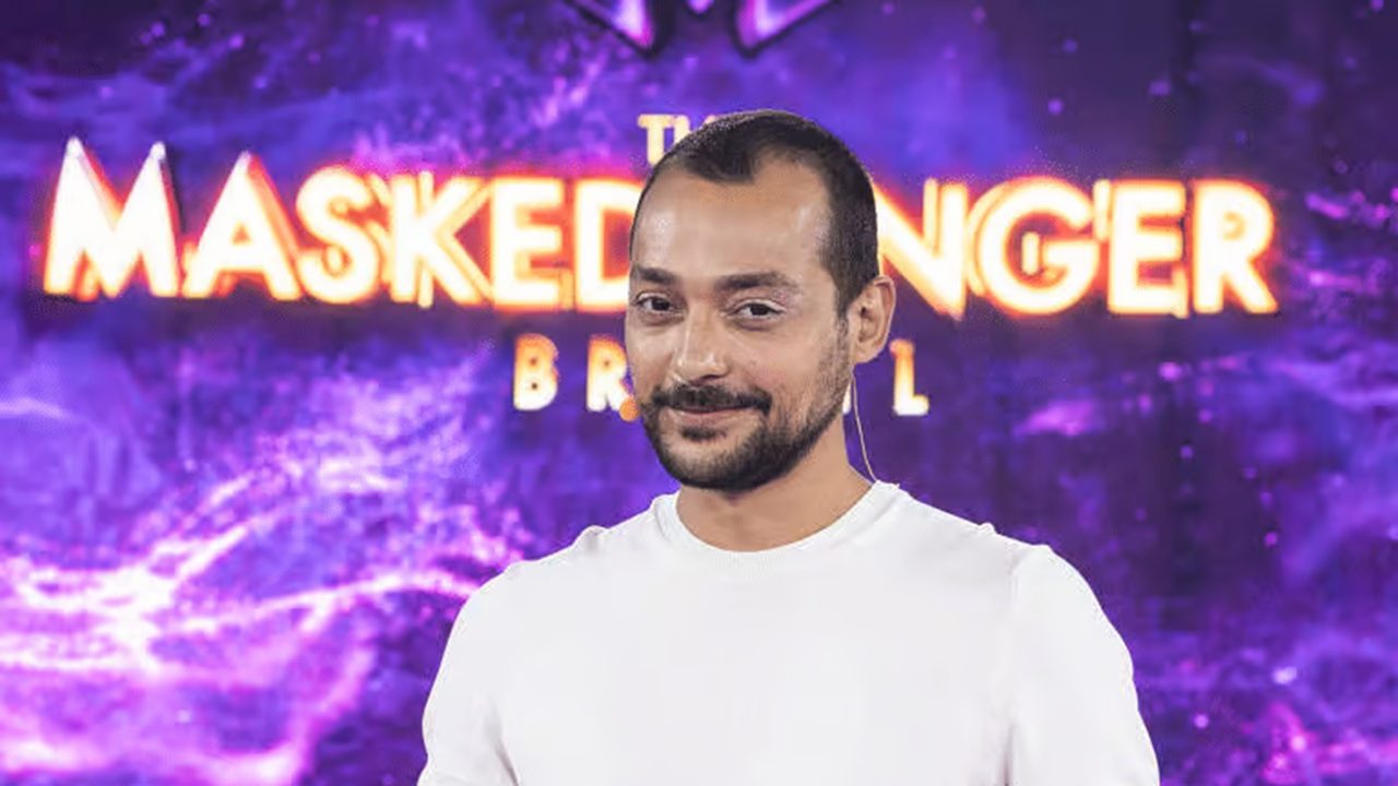 Homem sorrindo discretamente em frente a um painel roxo e iluminado do programa “The Masked Singer Brasil”. Ele veste camiseta branca e utiliza microfone de lapela, enquanto o fundo vibrante com luzes e efeitos gráficos destaca o ambiente de estúdio televisivo.