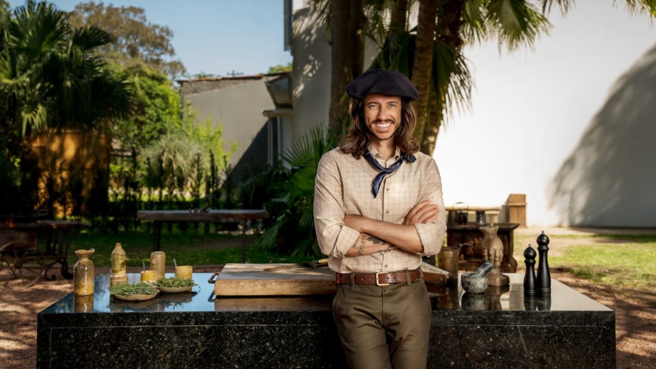 Homem de cabelos longos, usando boina e lenço no pescoço, sorri com os braços cruzados em frente a uma bancada de cozinha ao ar livre, com utensílios e ingredientes sobre a mesa e vegetação ao fundo