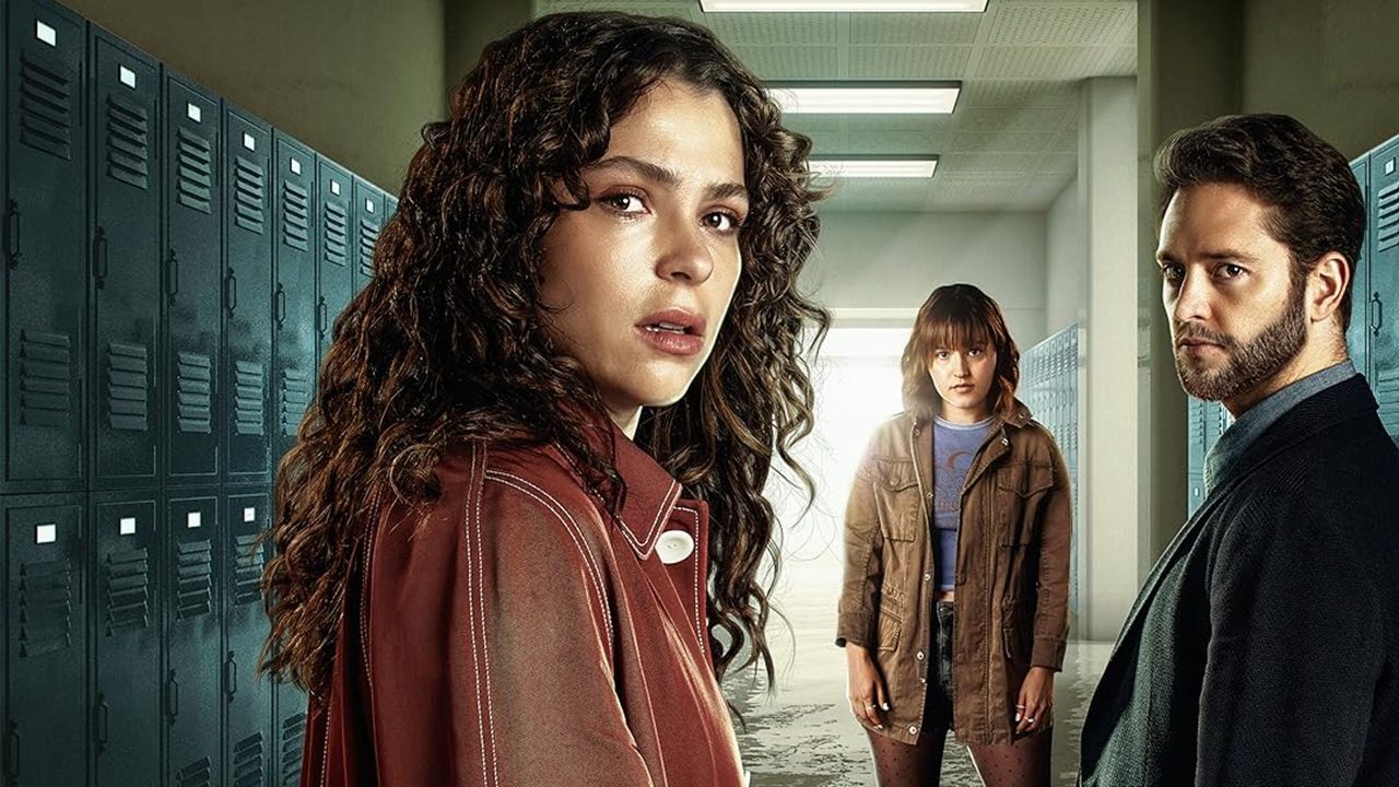 Três pessoas estão em um corredor de escola cercado por armários metálicos, com expressões sérias e tensas; a mulher à frente usa casaco marrom e olha fixamente para a câmera.