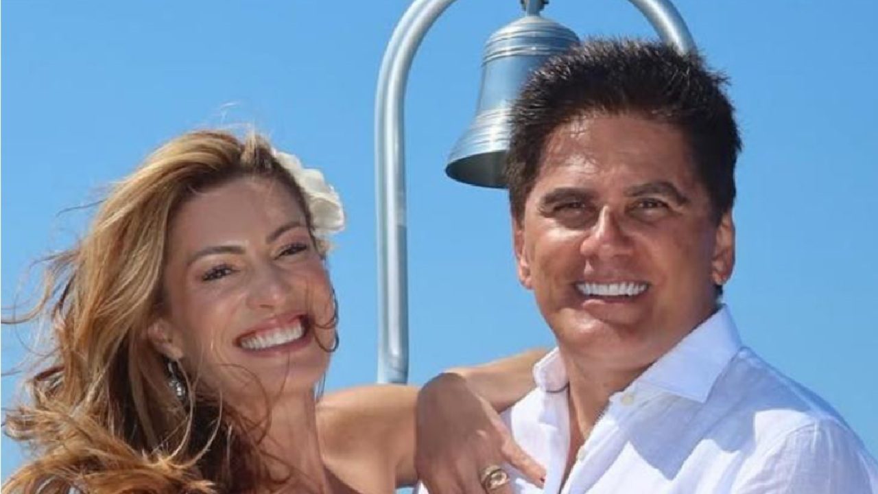 Casal sorridente posa ao ar livre em um dia ensolarado, com o céu azul ao fundo e um sino metálico atrás deles. A mulher usa flor no cabelo e o homem veste camisa branca, transmitindo leveza e alegria em um clima festivo ou de celebração