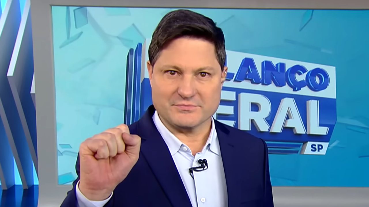 Foto do apresentador Eleandro Passaia no estúdio do telejornal Balanço Geral, usando um terno preto e blusa social azul