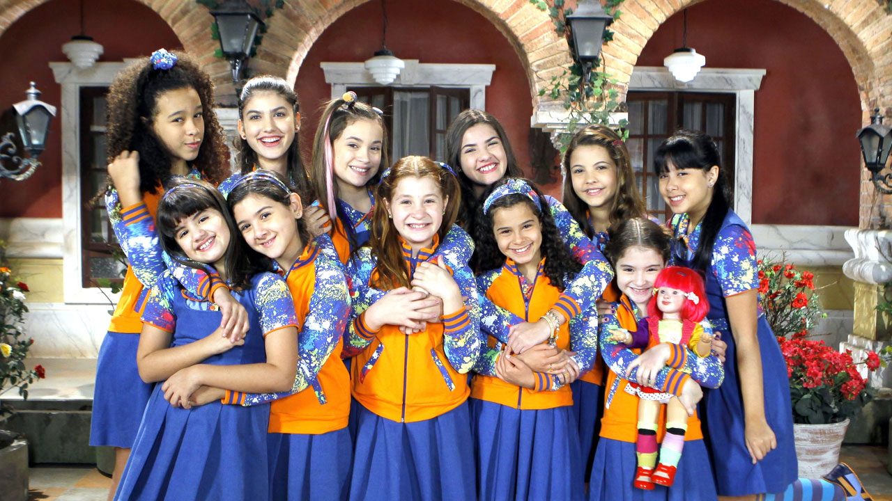 Crianças do elenco de Chiquititas com uniforme azul e laranja sorrindo abraçadas no pátio do orfanato Raio de Luz