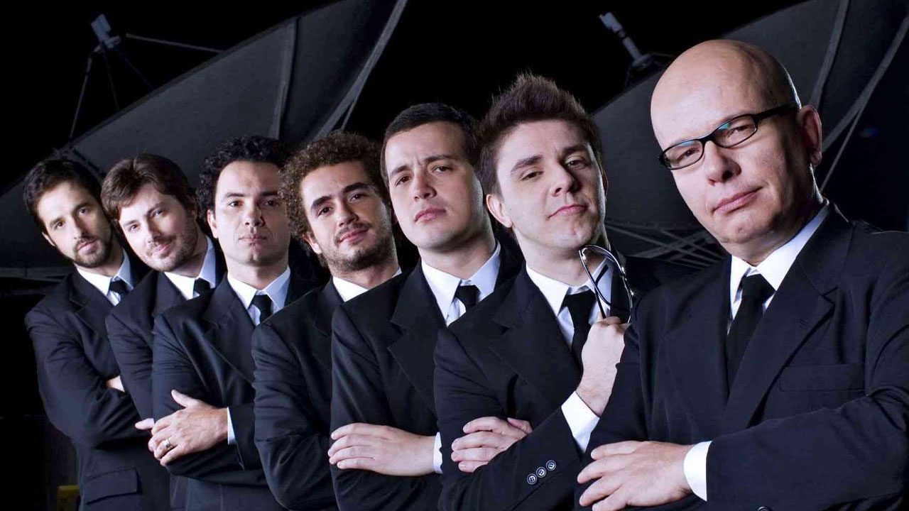 Elenco de apresentadores do CQC da Band em fila, de terno preto, braços cruzados, com fundo de antenas parabólicas