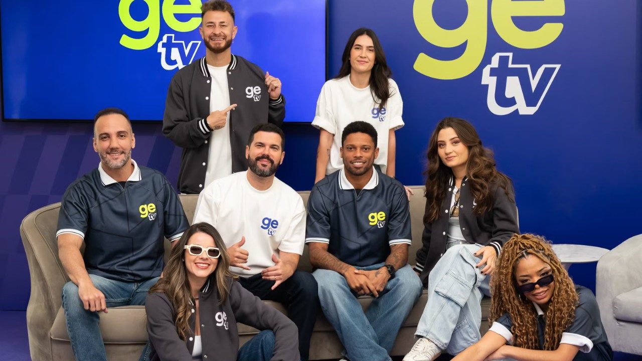 Elenco de talentos da GE TV posa em estúdio diante de telas com o logotipo do canal usando camisetas e jaquetas da marca