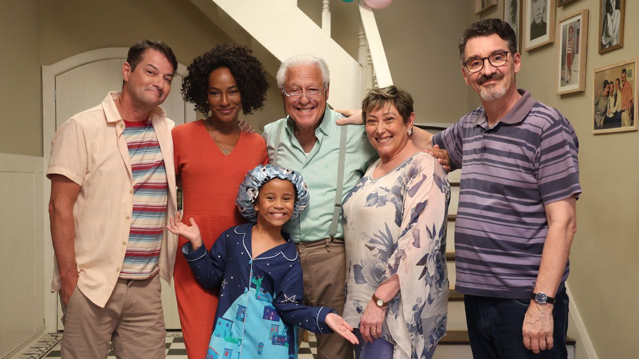 Elenco da série Mundo da Lua reunido em cenário de casa com escada e fotos na parede ao fundo