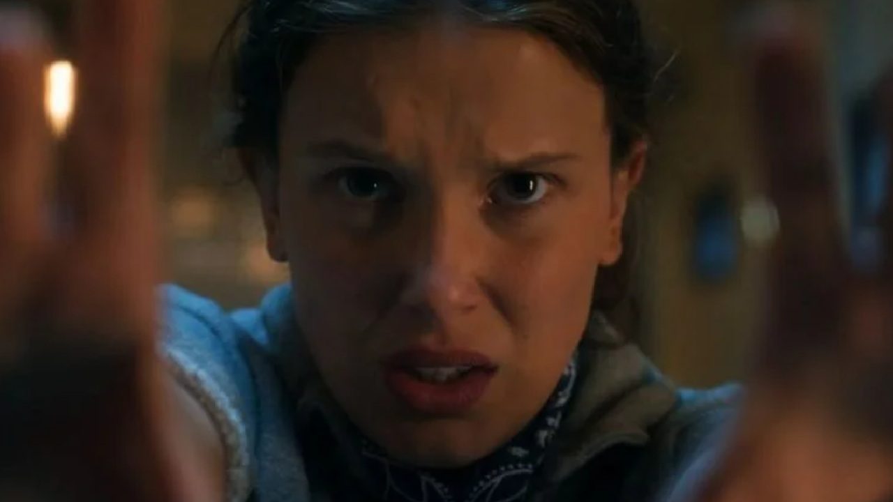 Adolescente olha fixamente para a frente com expressão tensa e concentrada, mantendo as mãos erguidas como se estivesse empurrando algo ou usando força intensa. A cena tem iluminação baixa e clima dramático, sugerindo momento de grande esforço ou tensão emocional