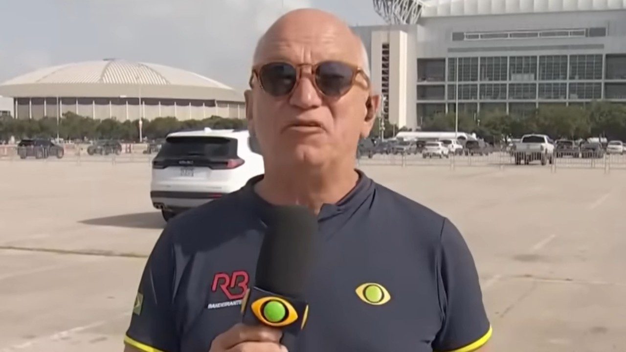 Elia Júnior de óculos escuros com microfone em estacionamento diante de arena esportiva
