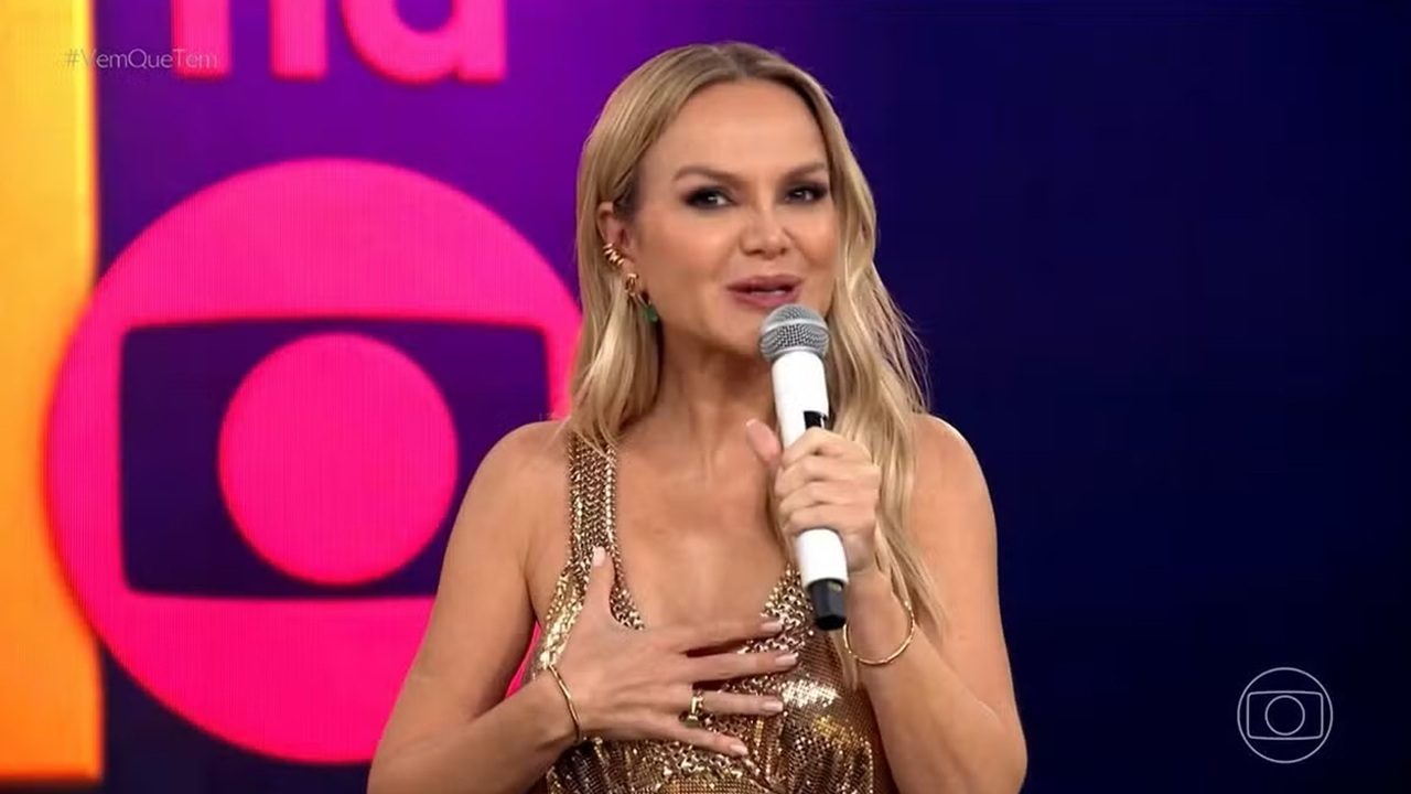 Apresentadora com vestido dourado fala ao microfone em palco da TV Globo com fundo colorido e logotipo da emissora