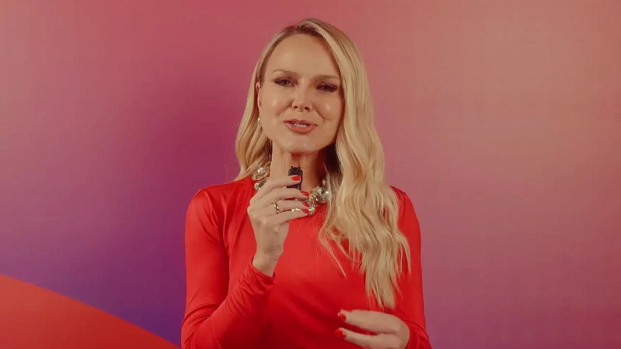 Mulher loira usando um vestido vermelho, segurando um pequeno microfone e falando para a câmera diante de um fundo colorido em tons de rosa e roxo.