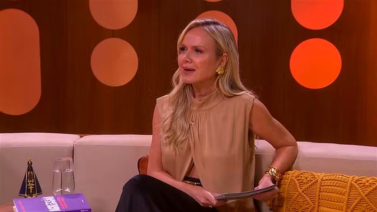 Apresentadora loira sentada em sofá fala durante programa de TV com cenário laranja ao fundo