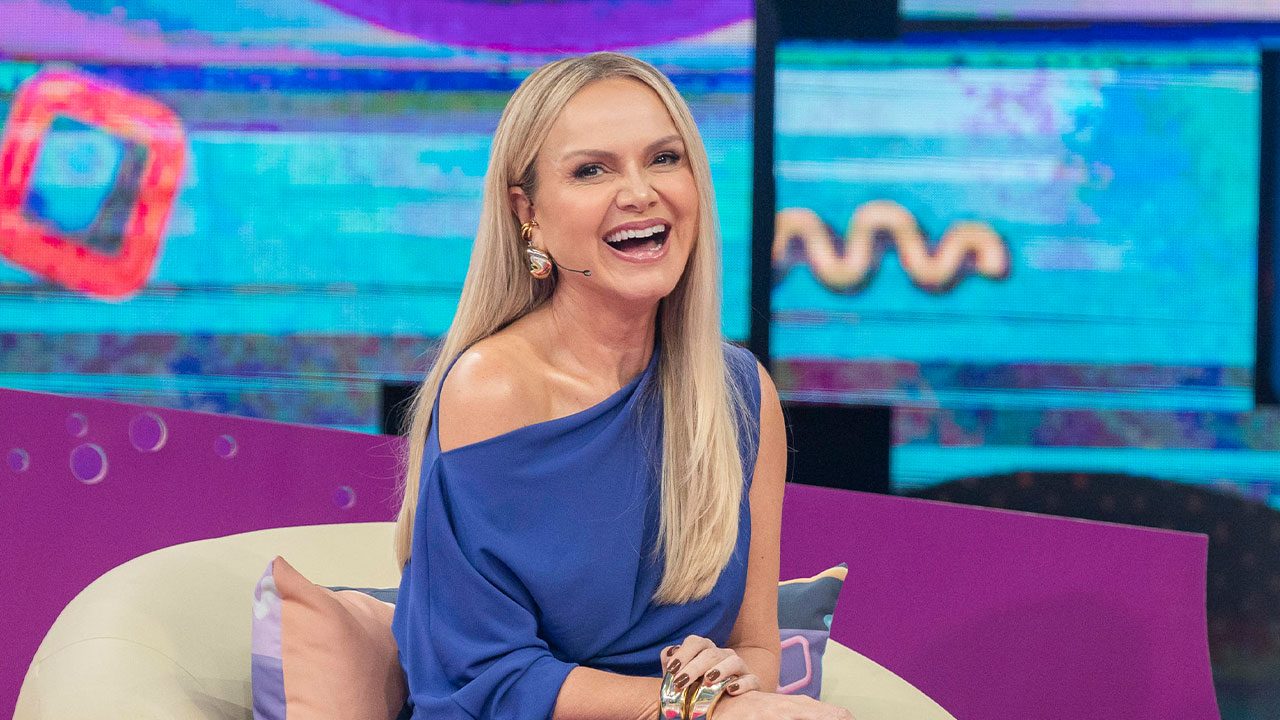 Eliana sentada e sorrindo com vestido azul em cenário colorido do Caldeirão com Mion